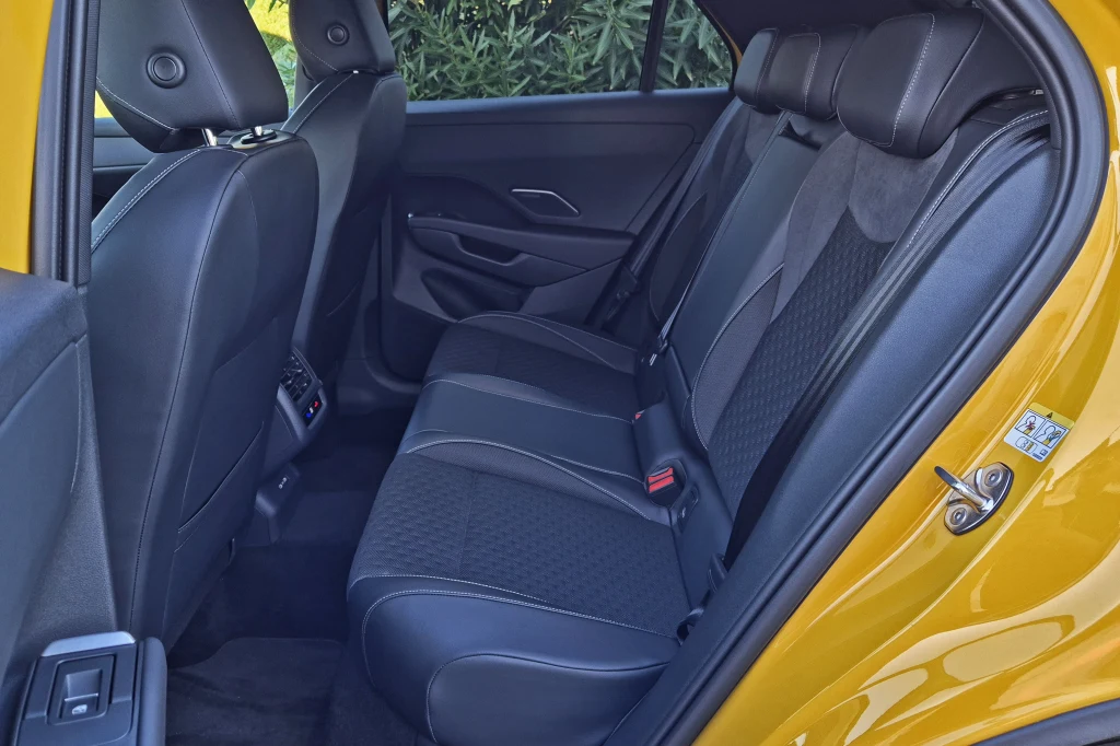 Ilość miejsca na tylnej kanapie jest wyraźnie większa niż w pierwszym T-Rocu. Nadal jednak przestrzeń ogranicza wysoki środkowy tunel Wnętrze samochodu z widokiem na tylną kanapę, tapicerka wykonana z czarnej tkaniny ze wstawkami materiałowymi, żółte drzwi boczne otwarte, na zewnątrz widoczne liście krzewu.