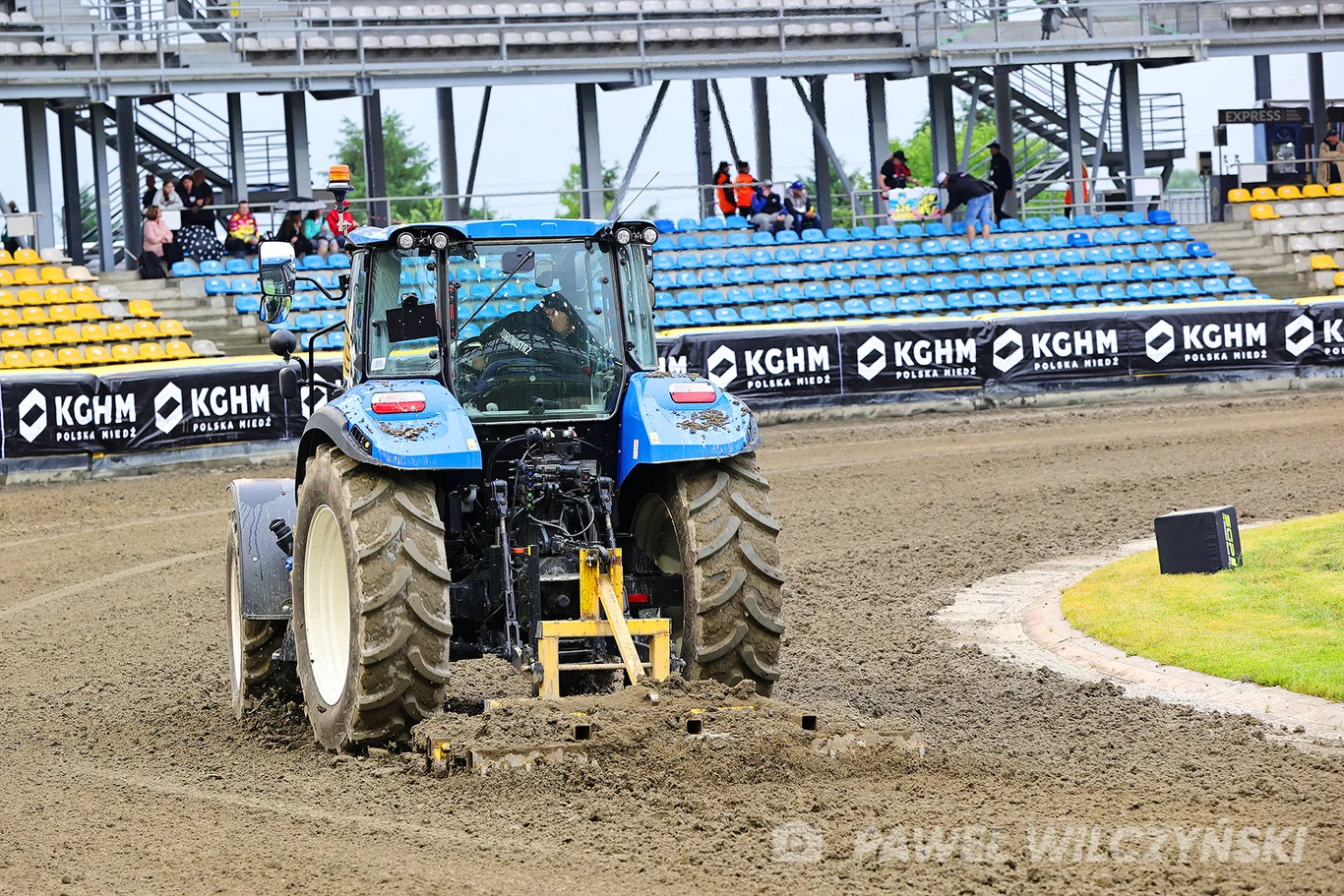 Niebieski traktor wyrównujący nawierzchnię toru żużlowego na stadionie z pustymi i częściowo zajętymi trybunami w tle. Po bokach widoczne są reklamy sponsorów, a obok toru kawałek zielonej murawy.
