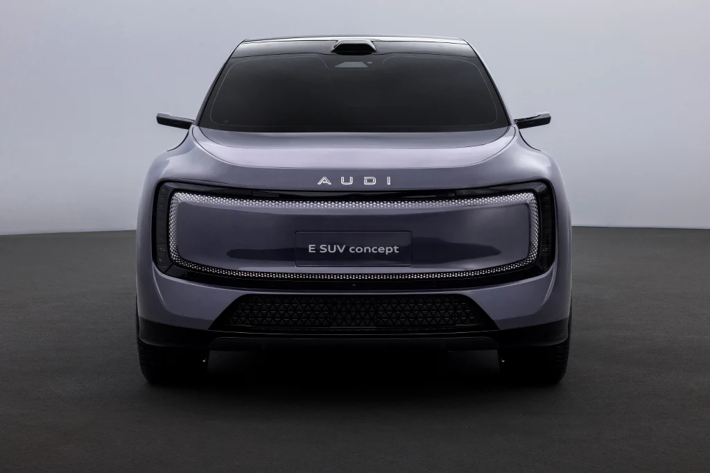 Futurystyczny elektryczny SUV marki Audi w kolorze szarym, prezentowany od frontu na neutralnym tle, z charakterystycznym zamkniętym grillem i nowoczesnym designem przednich świateł.