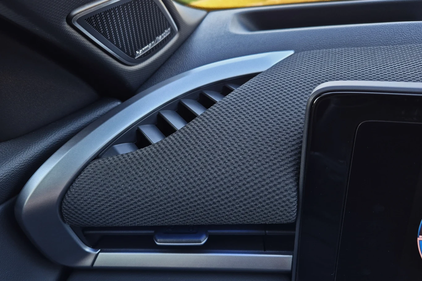 Pokryta tkaniną górna część deski rozdzielczej ma nadać wnętrzu T-Roca trochę szlachetności Fragment deski rozdzielczej samochodu z widocznym głośnikiem, kratką nawiewu oraz elementami tapicerowanymi i plastikowymi, obok części ekranu dotykowego.