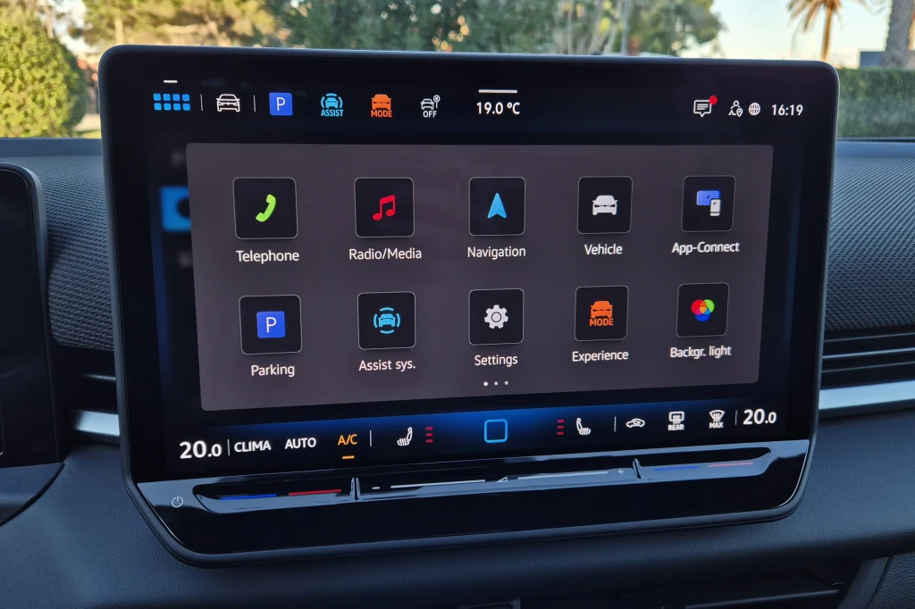 Centralny ekran ma przekątną 12,9 cala i dobrze już znane pola dotykowe do regulacji temperatury oraz głośności Centralny ekran dotykowy samochodu z wyświetlonym menu głównym systemu multimedialnego, pokazującym aplikacje takie jak telefon, radio, nawigacja, ustawienia oraz informacje o klimatyzacji w dolnej części ekranu.