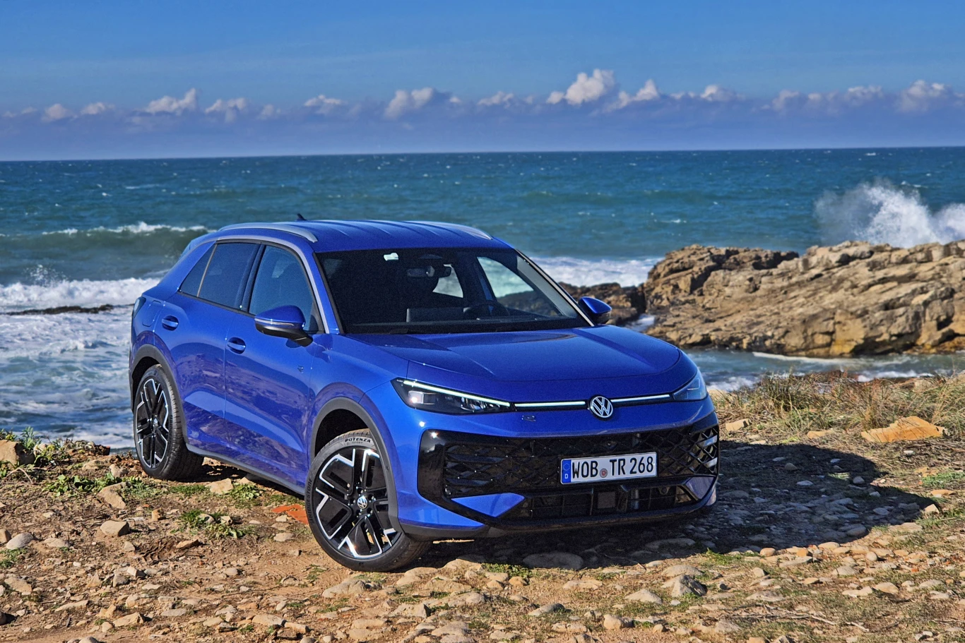 Volkswagen T-Roc Niebieski samochód marki Volkswagen zaparkowany na skalistym wybrzeżu, w tle fale morskie oraz błękitne niebo z widocznymi chmurami.