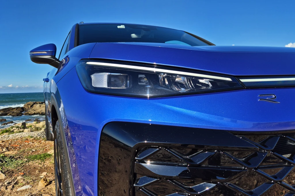 Volkswagen T-Roc Zbliżenie na przednią część niebieskiego samochodu osobowego, widoczny nowoczesny reflektor oraz charakterystyczna kratka wlotu powietrza, w tle wybrzeże i niebo.