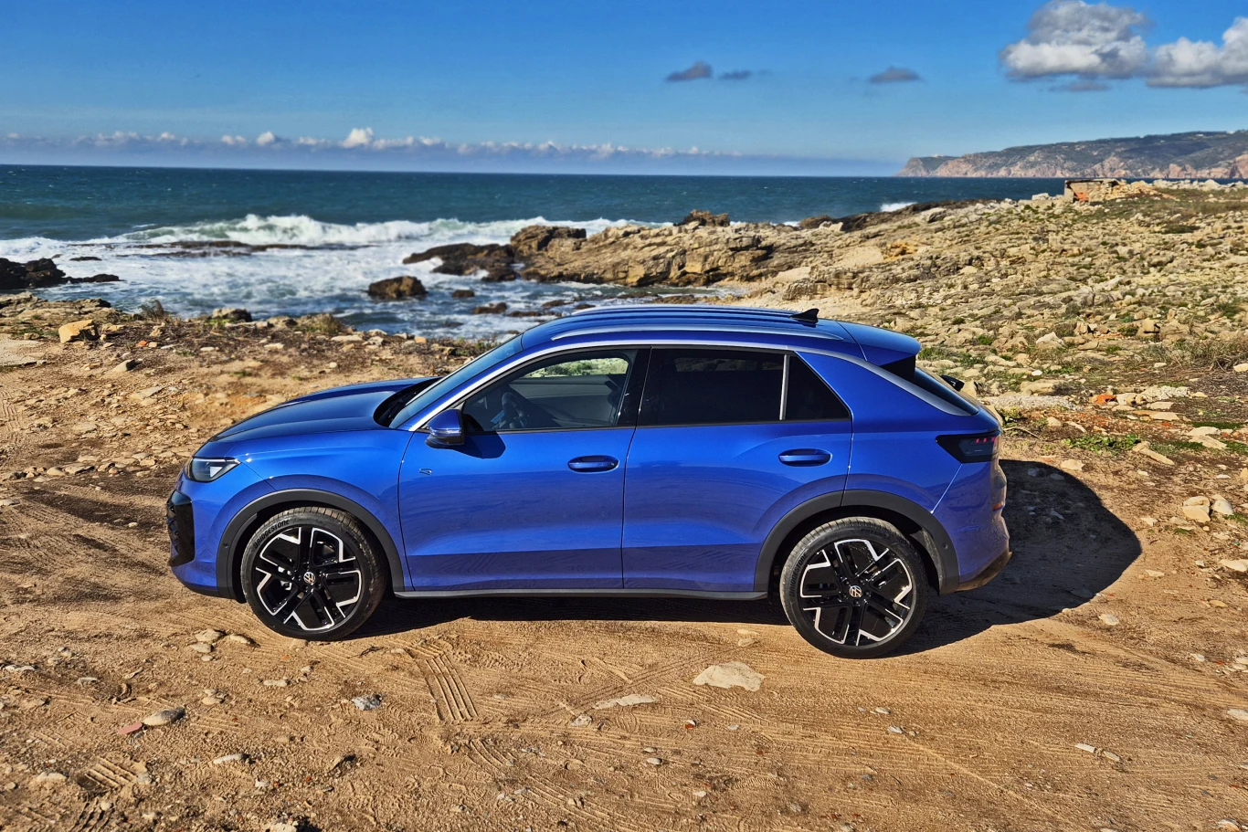 Volkswagen T-Roc Niebieski samochód SUV zaparkowany na kamienistym wybrzeżu w pobliżu oceanu, z falami rozbijającymi się o skały i rozległym widokiem na horyzont.