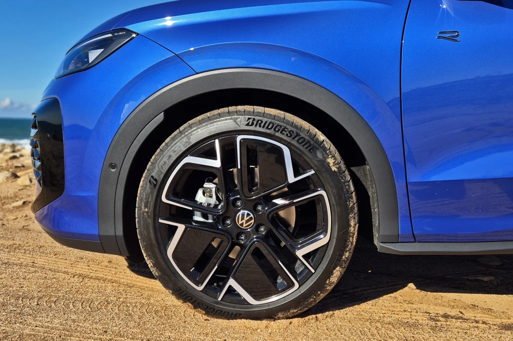 Volkswagen T-Roc Fragment niebieskiego samochodu z widocznym logotypem Volkswagen na feldze, czarne aluminiowe felgi z oponą marki Bridgestone, pojazd stoi na piaszczystym podłożu, w tle niewyraźne morze i kawałek nieba.