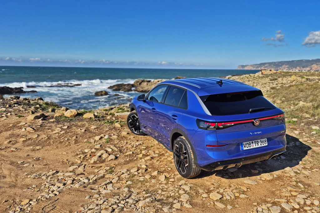 Volkswagen T-Roc Niebieski samochód stoi na kamienistej drodze przy wybrzeżu z widokiem na morze i skały, w tle rozciąga się błękitne niebo oraz urwiste wybrzeże.