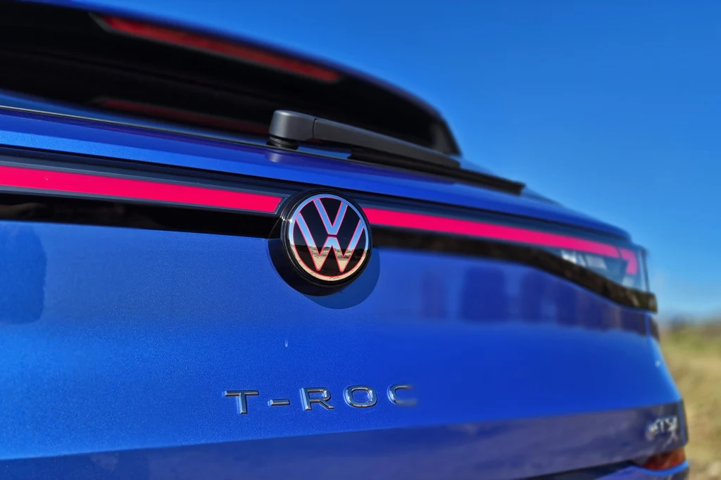 Volkswagen T-Roc Tył niebieskiego samochodu Volkswagen T-Roc z wyeksponowanym logo marki oraz czerwonym pasem oświetlenia na tle błękitnego nieba.