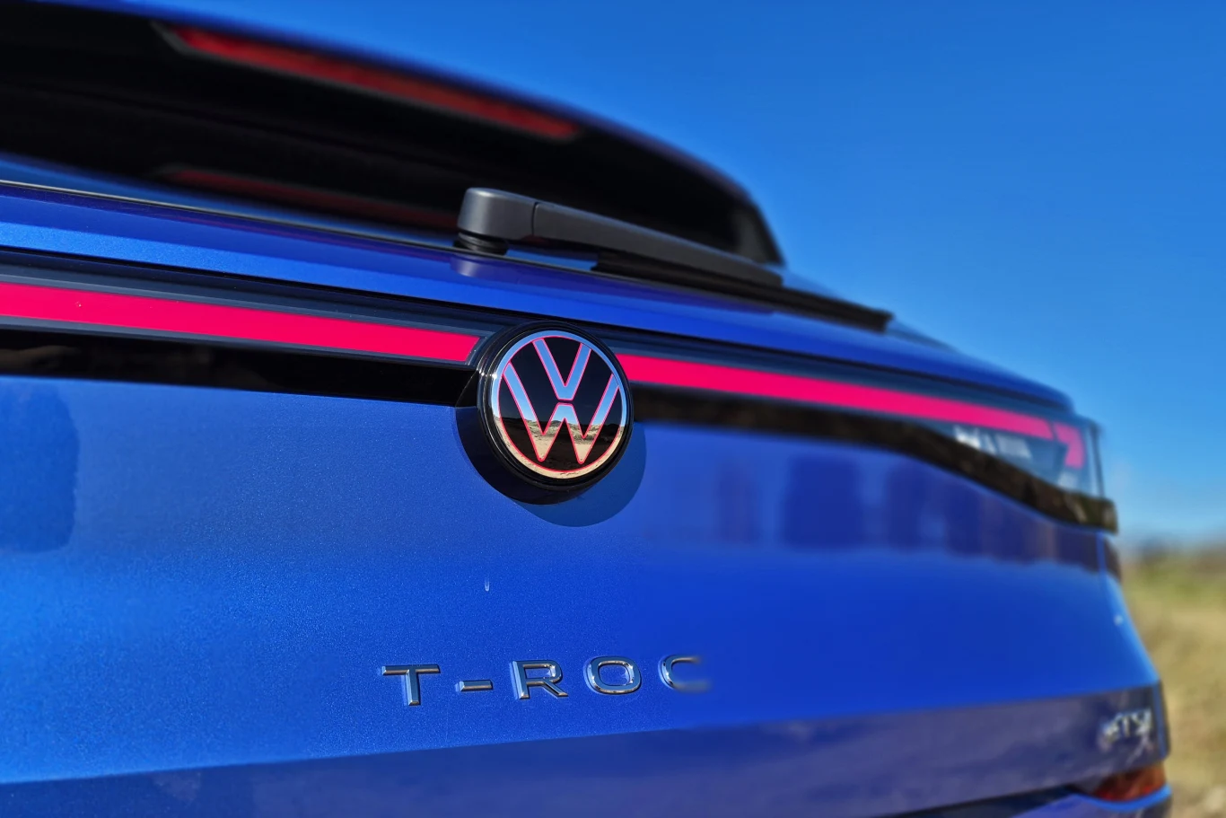 Volkswagen T-Roc Tył niebieskiego samochodu Volkswagen T-Roc z wyeksponowanym logo marki oraz czerwonym pasem oświetlenia na tle błękitnego nieba.