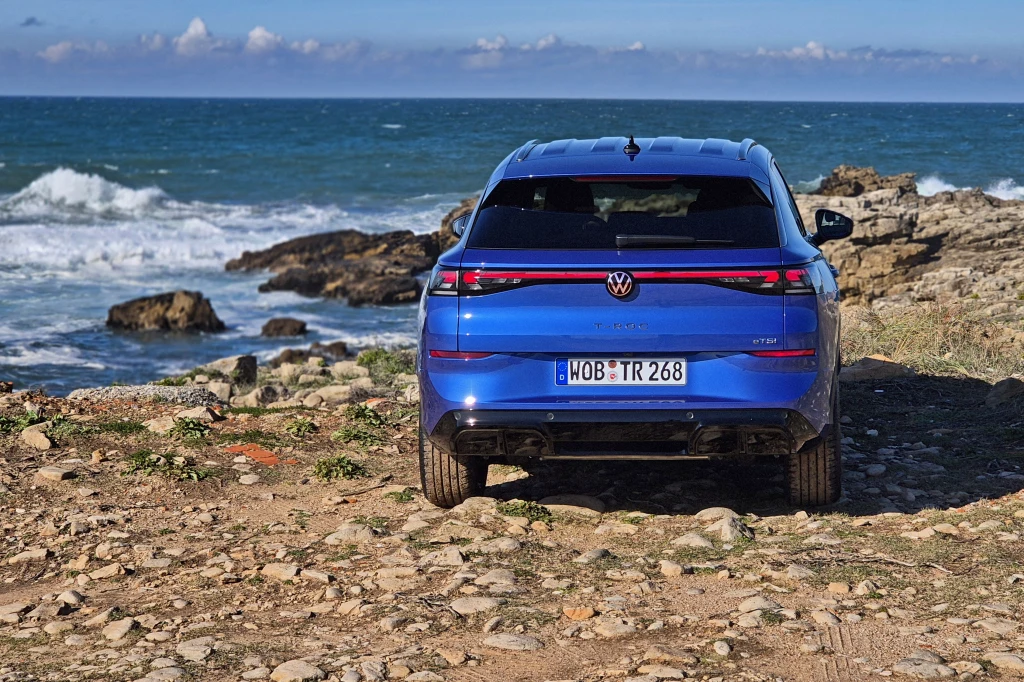 Volkswagen T-Roc Niebieski samochód marki Volkswagen ID.5 GTX zaparkowany tyłem przy skalistym wybrzeżu oceanu. Fale rozbijają się o brzeg, a wokół pojazdu widoczna jest kamienista droga oraz niskie rośliny.