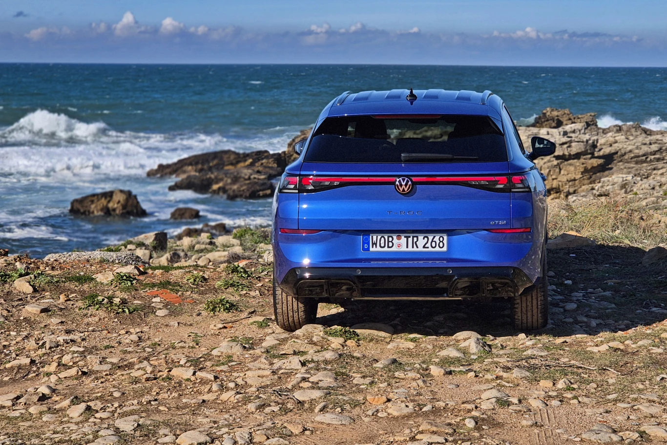Volkswagen T-Roc Niebieski samochód marki Volkswagen ID.5 GTX zaparkowany tyłem przy skalistym wybrzeżu oceanu. Fale rozbijają się o brzeg, a wokół pojazdu widoczna jest kamienista droga oraz niskie rośliny.