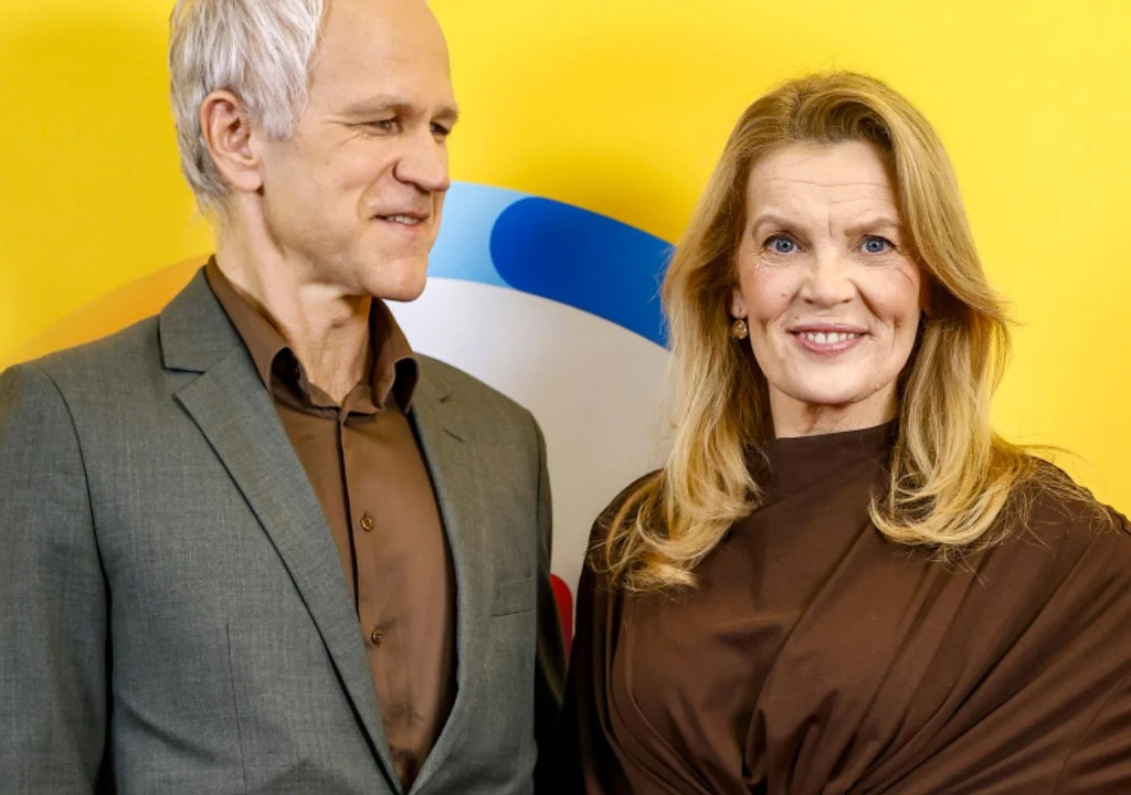 Dwoje dojrzałych ludzi stojących razem na tle żółtej ściany z kolorowymi elementami, oboje ubrani elegancko, kobieta uśmiechnięta, mężczyzna patrzy na nią.