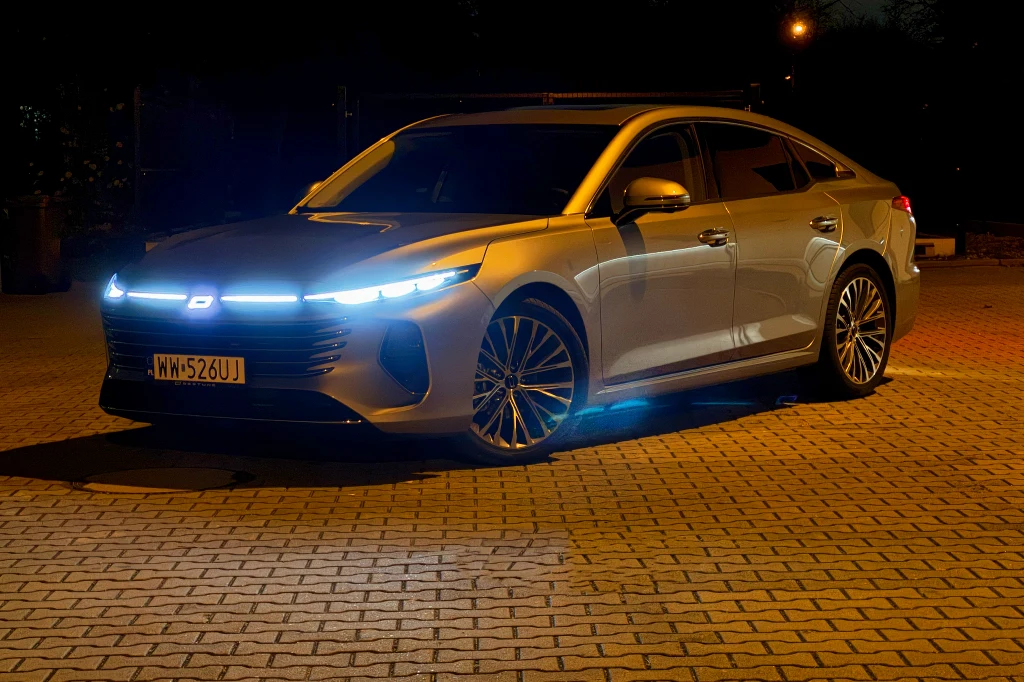 Srebrny nowoczesny samochód osobowy z podświetleniem LED i światłami do jazdy dziennej stoi na parkingu z kostki brukowej po zmroku.