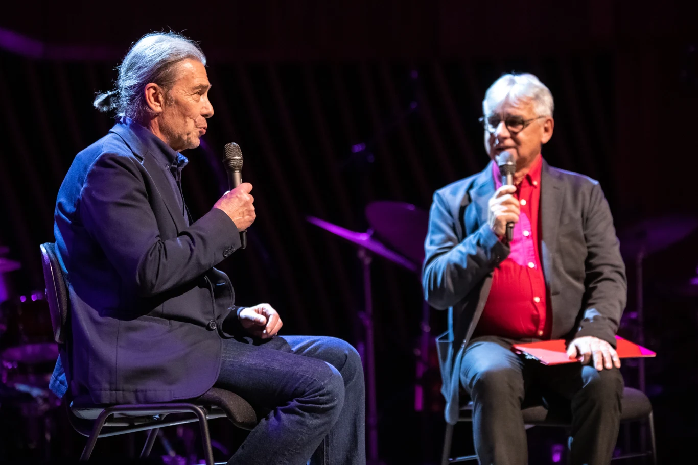 Dwaj mężczyźni siedzą na scenie, rozmawiając ze sobą z użyciem mikrofonów. Jeden z nich trzyma teczkę na kolanach, tło jest ciemne i sugeruje atmosferę wydarzenia lub wywiadu.