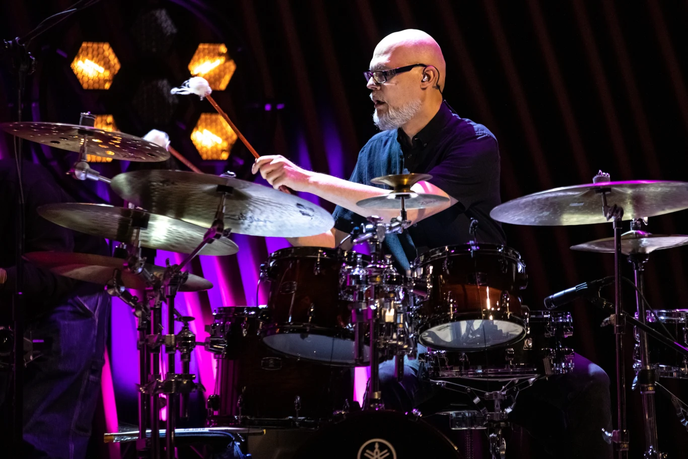Łysy mężczyzna w okularach gra na perkusji podczas koncertu, otoczony przez sceniczne światła i elementy zestawu perkusyjnego, w tle kolorowe iluminacje.
