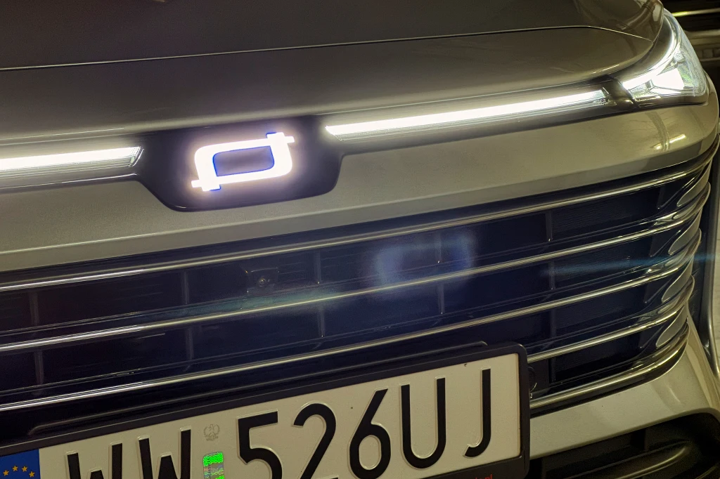 Zbliżenie na przednią część samochodu z podświetlonym logo marki oraz tablicą rejestracyjną samochodu z numerem WW 526UJ. Na grillu widoczne są poziome listwy, a maska odbija światło.