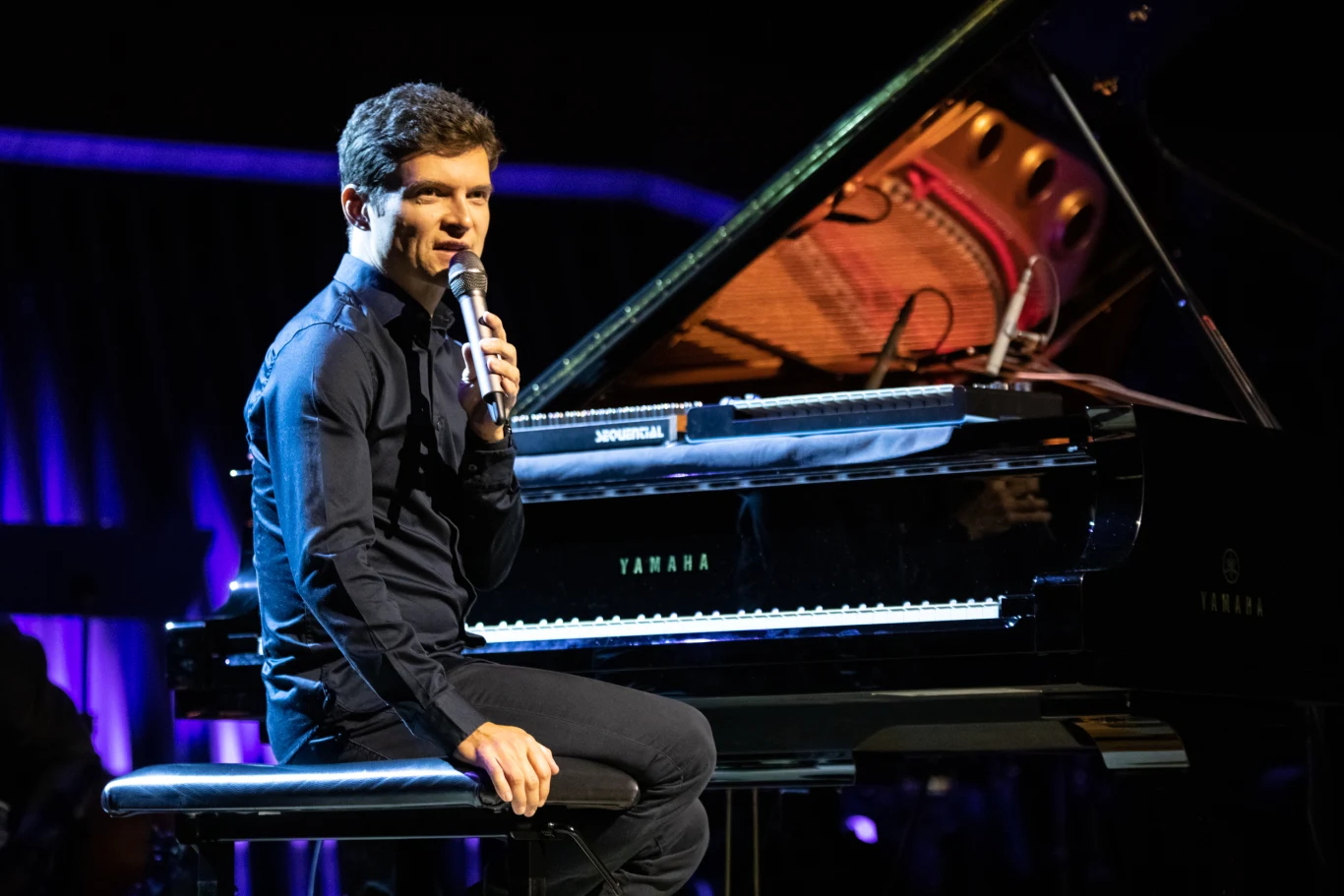 Mężczyzna w eleganckim, ciemnym ubraniu siedzi przy fortepianie Yamaha na scenie, trzyma mikrofon i patrzy w stronę publiczności, w tle widoczne kolorowe światła sceniczne.