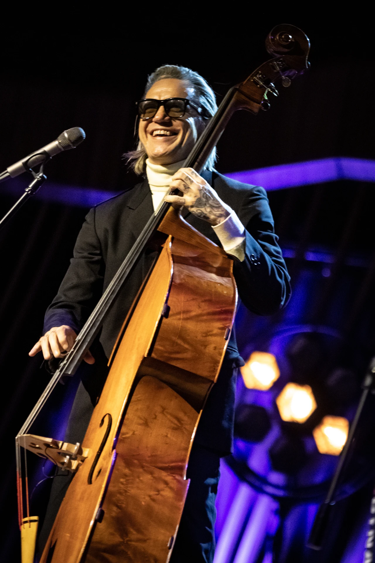 Mężczyzna w ciemnych okularach i garniturze gra na kontrabasie, stojąc na scenie oświetlonej ciepłym światłem, w tle widoczne reflektory i elementy scenografii.