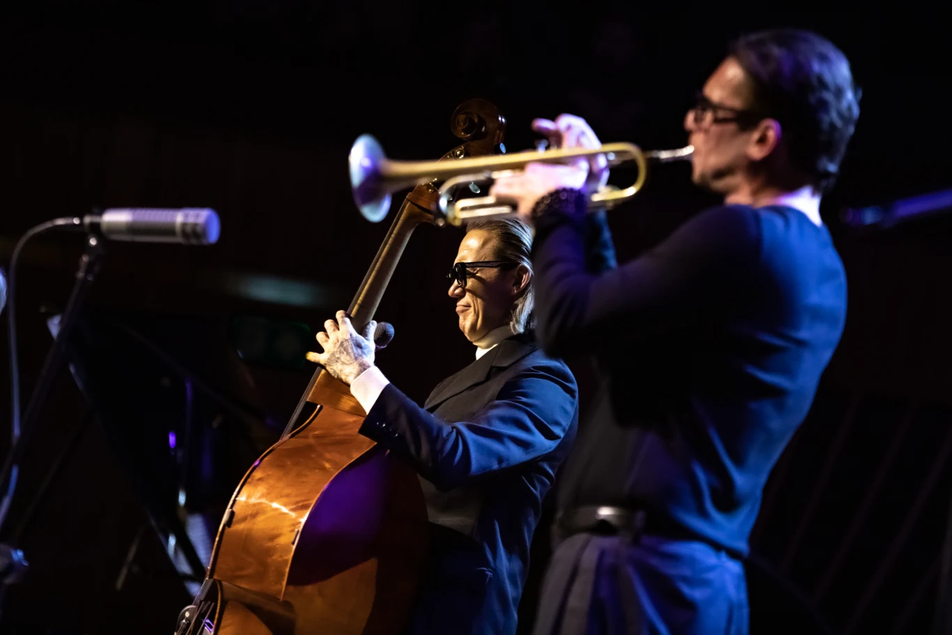 Dwaj muzycy podczas koncertu jazzowego, jeden gra na kontrabasie, drugi na trąbce. Artyści skoncentrowani na swoich instrumentach, w tle widoczny mikrofon oraz lekko rozmazane sylwetki. Na scenie panuje nastrojowe oświetlenie podkreślające atmosferę wy...