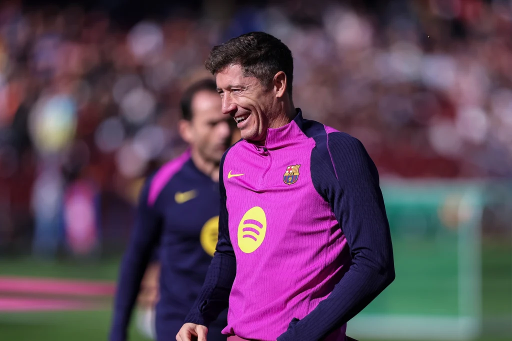 Robert Lewandowski Piłkarz w stroju treningowym FC Barcelony uśmiecha się podczas zajęć na boisku, w tle widoczny jest drugi zawodnik i rozmazane trybuny stadionu.