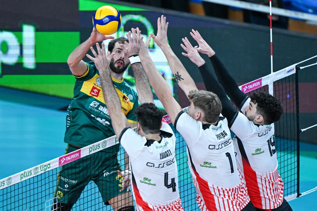 Zawodnik drużyny w zielonym stroju atakuje piłkę siatkową nad siatką, naprzeciwko trzech zawodników w biało-czerwonych koszulkach, którzy próbują zablokować uderzenie podczas meczu ligowego.
