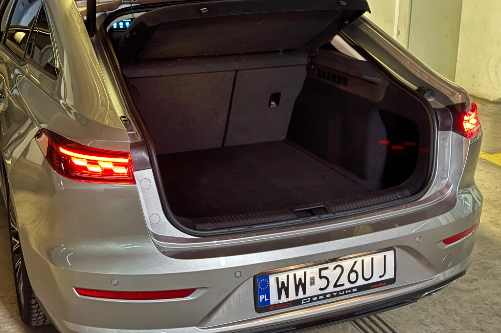 Srebrny samochód z otwartym bagażnikiem, tylne światła świecą się na czerwono, widoczna polska tablica rejestracyjna oraz oświetlone wnętrze garażu.