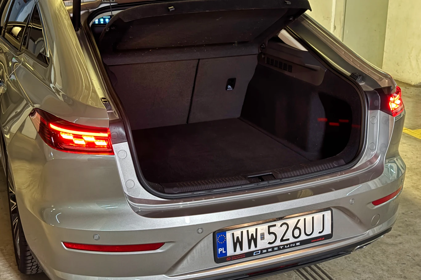 Srebrny samochód z otwartym bagażnikiem, tylne światła świecą się na czerwono, widoczna polska tablica rejestracyjna oraz oświetlone wnętrze garażu.