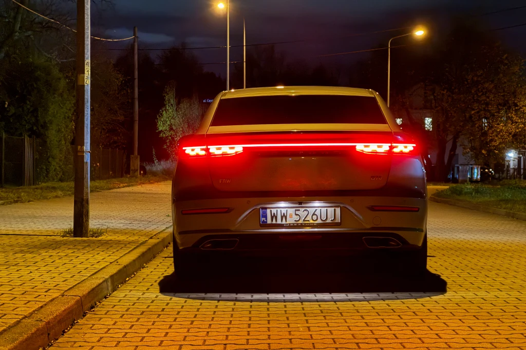 Samochód osobowy zaparkowany na ulicy nocą, tylne światła zapalone, tablica rejestracyjna widoczna, oświetlenie uliczne rozświetla brukowaną drogę, otoczenie z drzewami i domami w tle.