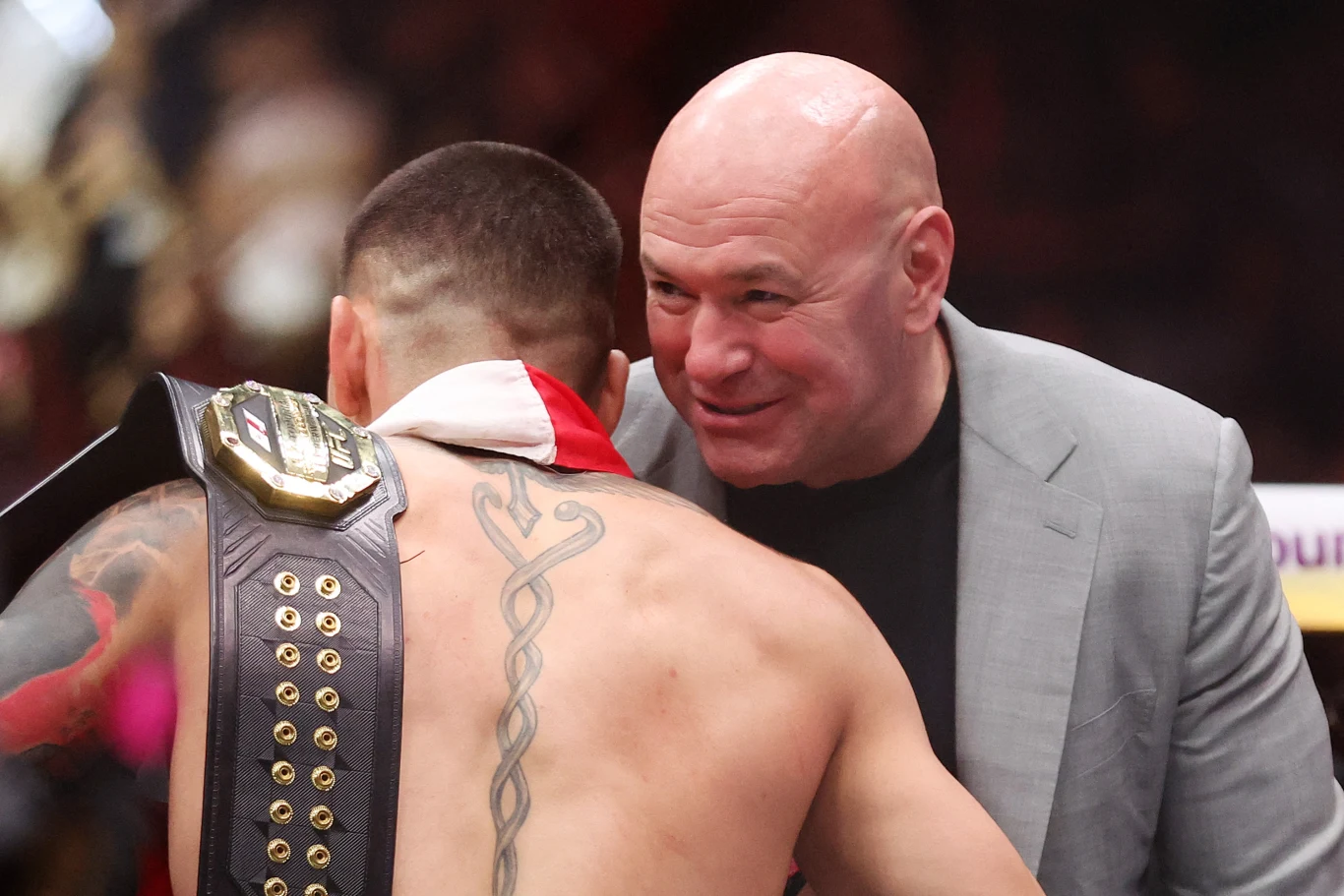 Dana White Mężczyzna z tatuażem na plecach i pasem mistrzowskim na ramieniu obejmuje drugiego mężczyznę w garniturze, który uśmiecha się i rozmawia z nim. Atmosfera sugeruje celebrację sportowego zwycięstwa.