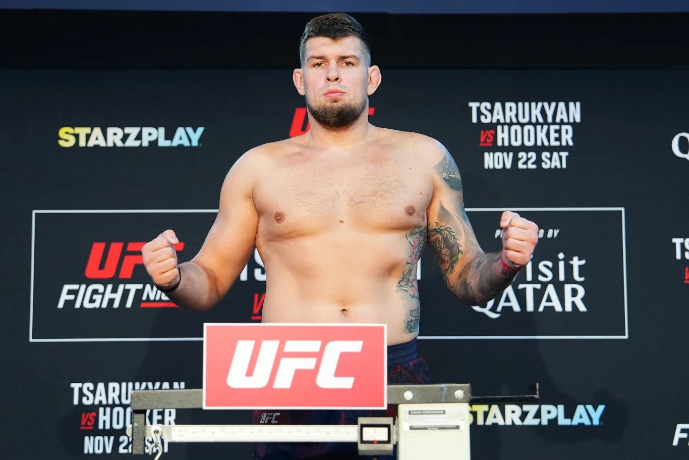 UFC: Marek Bujło Zawodnik MMA w krótkich spodenkach stoi na wadze podczas oficjalnego ważenia UFC, z zaciśniętymi pięściami po bokach, w tle widoczna jest tablica z informacjami o wydarzeniu i logo organizatorów.