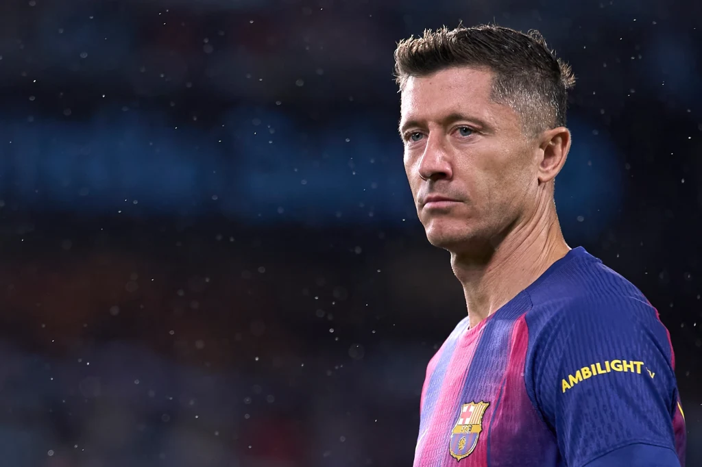 Robert Lewandowski Piłkarz w koszulce katalońskiego klubu FC Barcelona na tle nieostrego stadionu, skoncentrowany i lekko zwrócony bokiem, widoczne krótkie ciemne włosy i charakterystyczna mina skupienia.