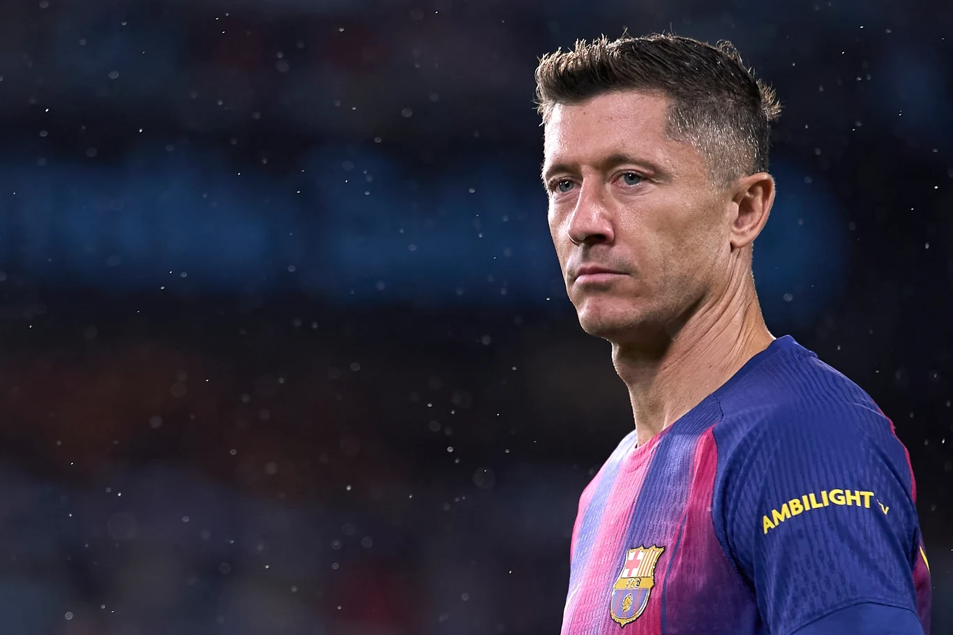 Robert Lewandowski Piłkarz w koszulce katalońskiego klubu FC Barcelona na tle nieostrego stadionu, skoncentrowany i lekko zwrócony bokiem, widoczne krótkie ciemne włosy i charakterystyczna mina skupienia.