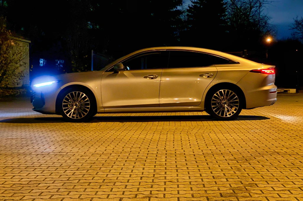 Srebrny sedan o nowoczesnym designie stoi zaparkowany na brukowanym placu, reflektory samochodu są włączone. W tle widoczne są drzewa oraz delikatne oświetlenie uliczne, co sugeruje wieczorną porę.