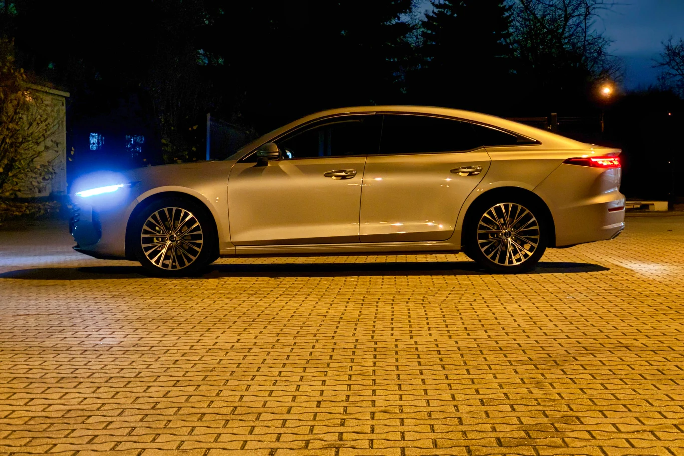 Srebrny sedan o nowoczesnym designie stoi zaparkowany na brukowanym placu, reflektory samochodu są włączone. W tle widoczne są drzewa oraz delikatne oświetlenie uliczne, co sugeruje wieczorną porę.