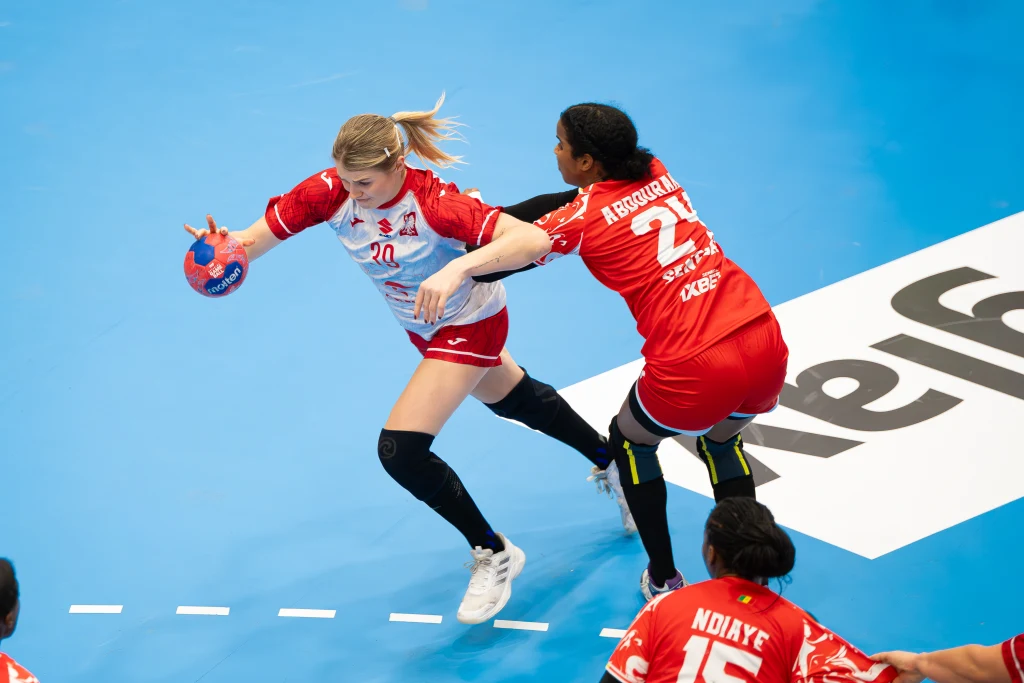 Intensywna akcja podczas meczu piłki ręcznej kobiet, zawodniczka w białym stroju stara się przejść obok rywalki w czerwonym stroju, mając piłkę w ręku, obie walczą o przewagę na niebieskim boisku.