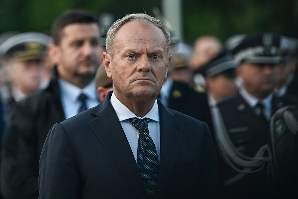 Donald Tusk odniósł się do żądań premiera Węgier Viktora Orbana w sprawie Ukrainy Mężczyzna w garniturze i krawacie stoi na pierwszym planie, w tle widoczni są umundurowani mężczyźni, prawdopodobnie przedstawiciele służb mundurowych, scena odbywa się na zewnątrz.