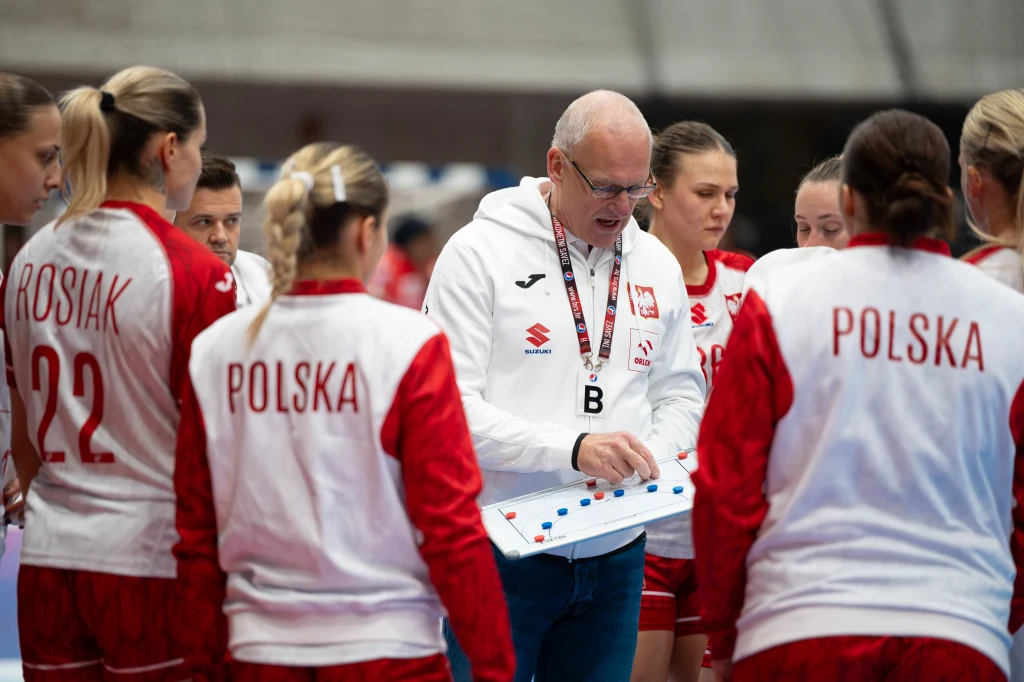 Trener w białej bluzie analizuje taktykę z zawodniczkami polskiej drużyny piłki ręcznej, ubrane w czerwono-białe stroje z napisem Polska na plecach, skupione na tablicy z rysunkiem schematu gry.