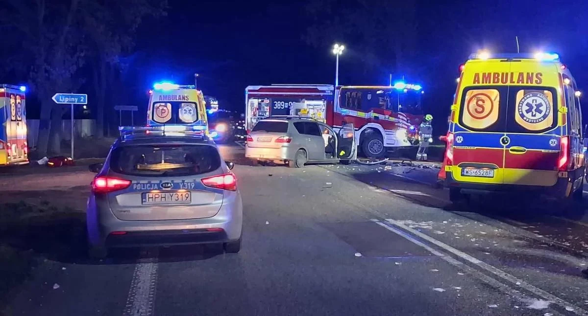 Na drodze krajowej nr 2 w miejscowości Ługi-Rętki (woj. mazowieckie) doszło do zderzenia dwóch pojazdów osobowych. Nocna akcja ratunkowa po wypadku drogowym z udziałem kilku pojazdów, wokół których stoją radiowóz policyjny, dwa ambulanse i wóz strażacki. Silne światła alarmowe rozświetlają ciemną jezdnię, na której widoczne są fragmenty rozbitych samochodów.