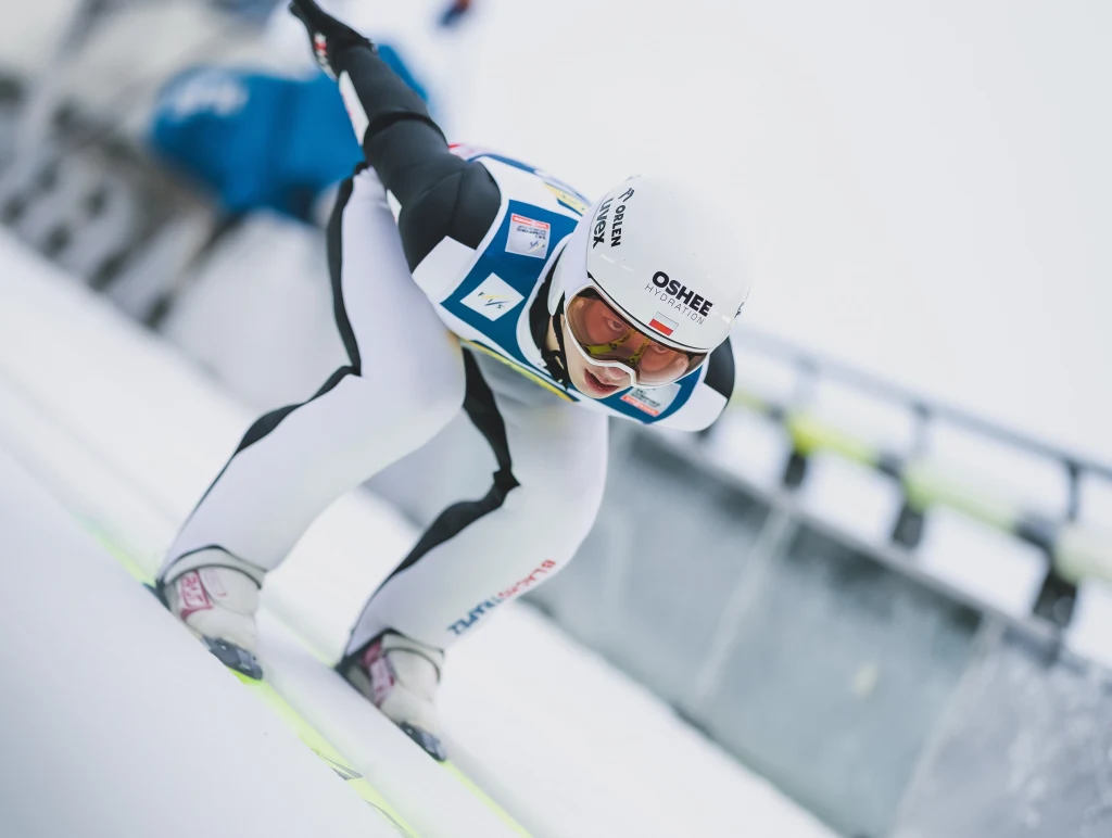 Kacper Tomasiak Zawodnik w białym kombinezonie i kasku podczas zjazdu na skoczni narciarskiej w trakcie skoku, dynamiczna poza i intensywna koncentracja, w tle elementy infrastruktury sportowej.