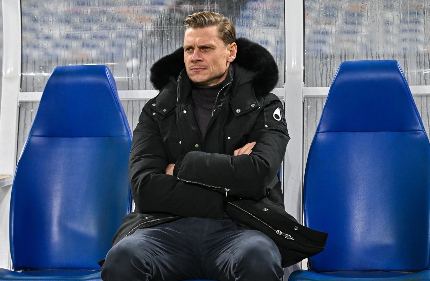 Łukasz Piszczek Mężczyzna w czarnej kurtce z futrzanym kołnierzem siedzi ze skrzyżowanymi rękami na niebieskim fotelu stadionowym, w tle rozmyte krzesełka trybun.