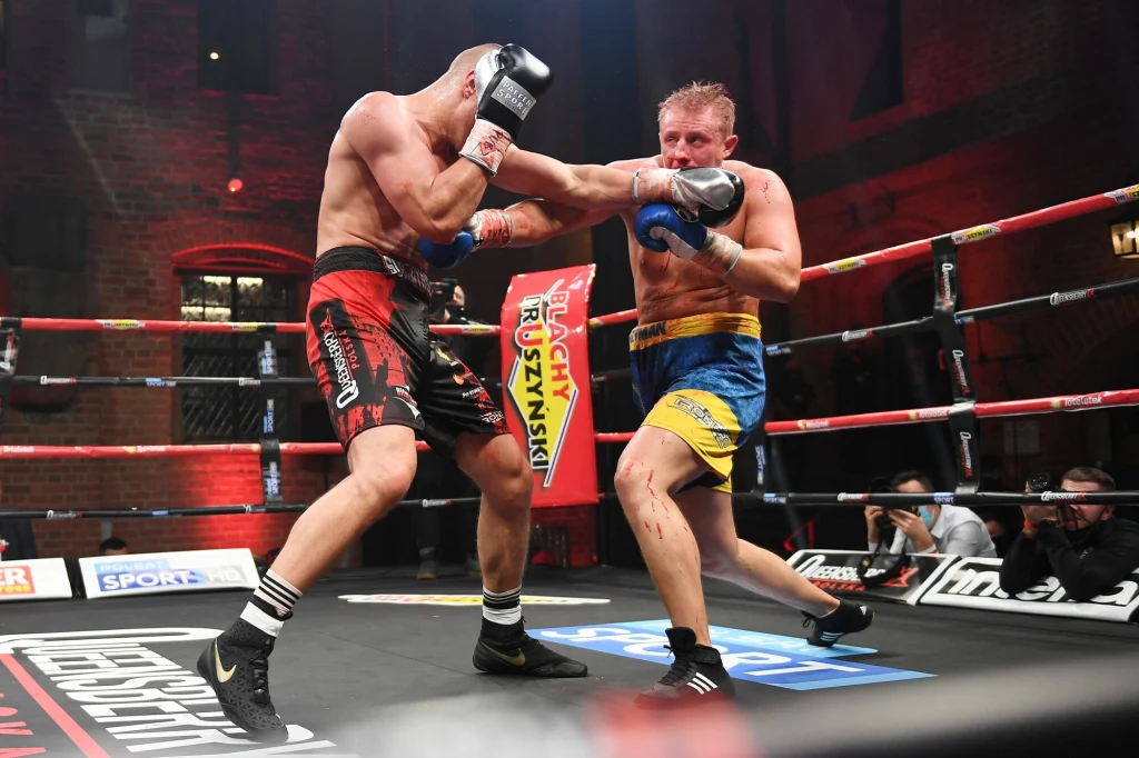 Dwóch bokserów walczących w ringu, jeden z zawodników trafia przeciwnika lewym prostym w twarz, otoczeni są linami ringu i obserwowani przez sędziów oraz widzów, tło stanowią ceglane ściany hali.