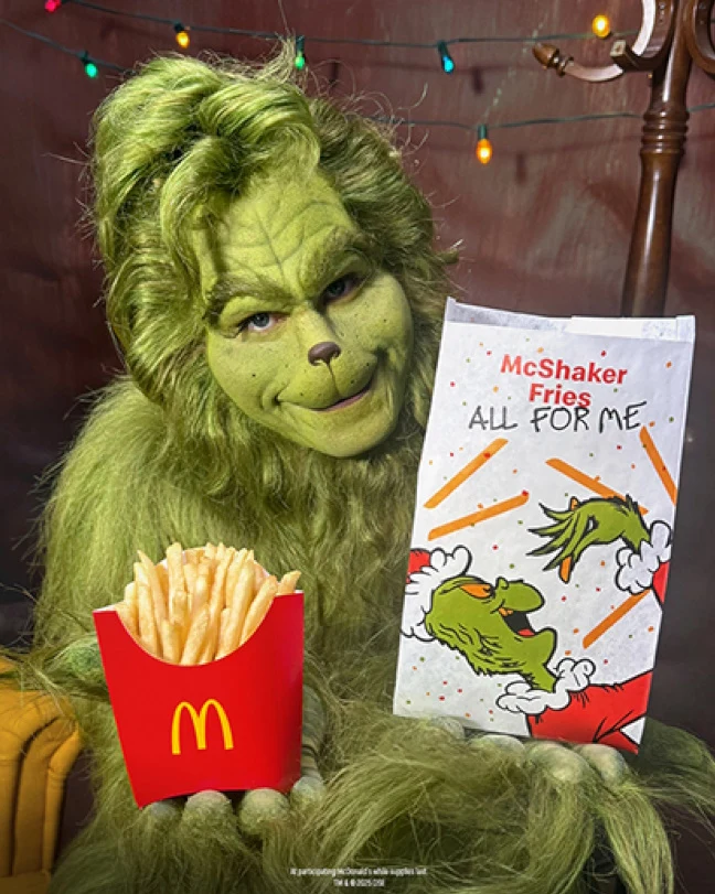 Postać ucharakteryzowana na Grincha siedzi na tle świątecznych lampek, trzymając opakowanie frytek McDonald's oraz papierową torbę z ilustracją Grincha i napisem McShaker Fries All For Me.