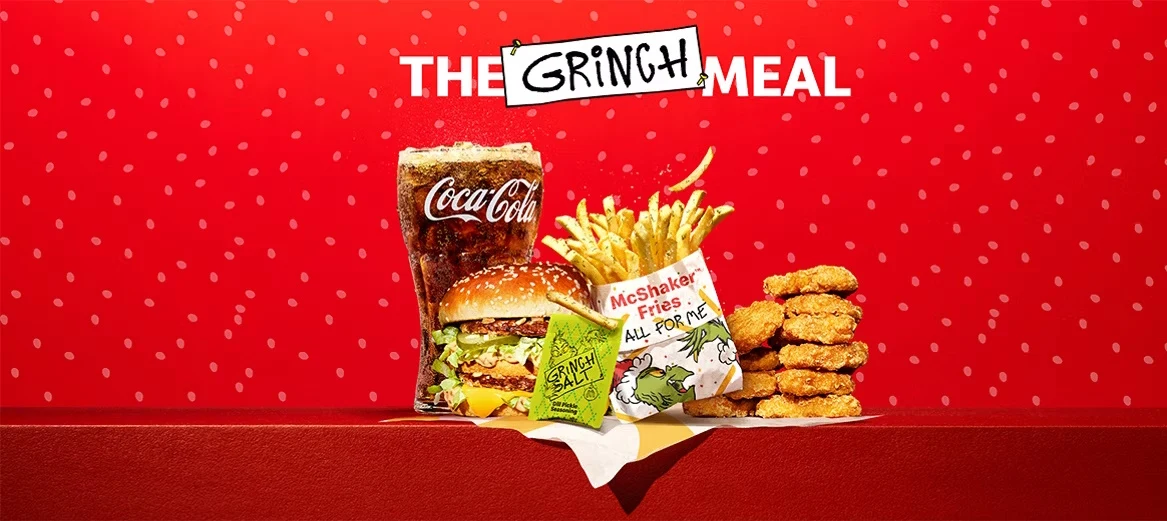 Zestaw Grinch Meal składający się z dużego napoju Coca-Cola, frytek, burgera z sałatą i dodatkami, porcji nuggetsów oraz zielonego sosu na tle czerwonej tapety z drobnym wzorem i zabawnym napisem.