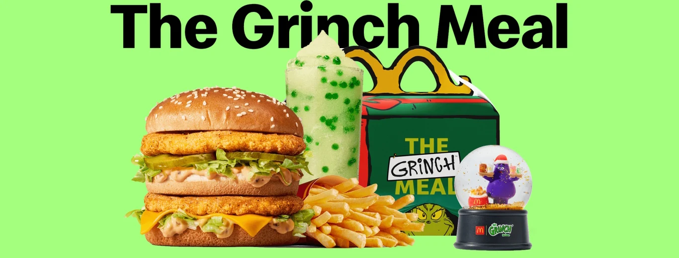 Zestaw jedzenia składający się z podwójnego burgera z kurczakiem, porcji frytek, zielonego deseru w kubku z bitą śmietaną oraz kolorowego pudełka z napisem The Grinch Meal, obok którego znajduje się ozdobna kula z postacią z bajki.