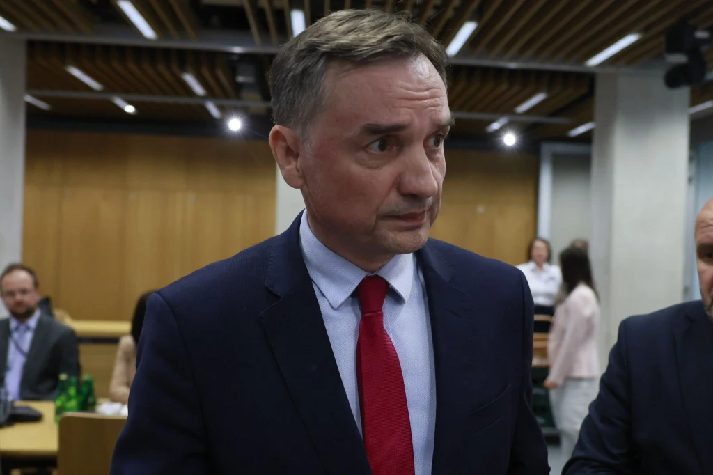 Zbigniew Ziobro Mężczyzna w garniturze i czerwonym krawacie stoi we wnętrzu nowoczesnej sali konferencyjnej, w tle widoczne są inne osoby siedzące przy stołach oraz elementy architektury z drewnianym sufitem.