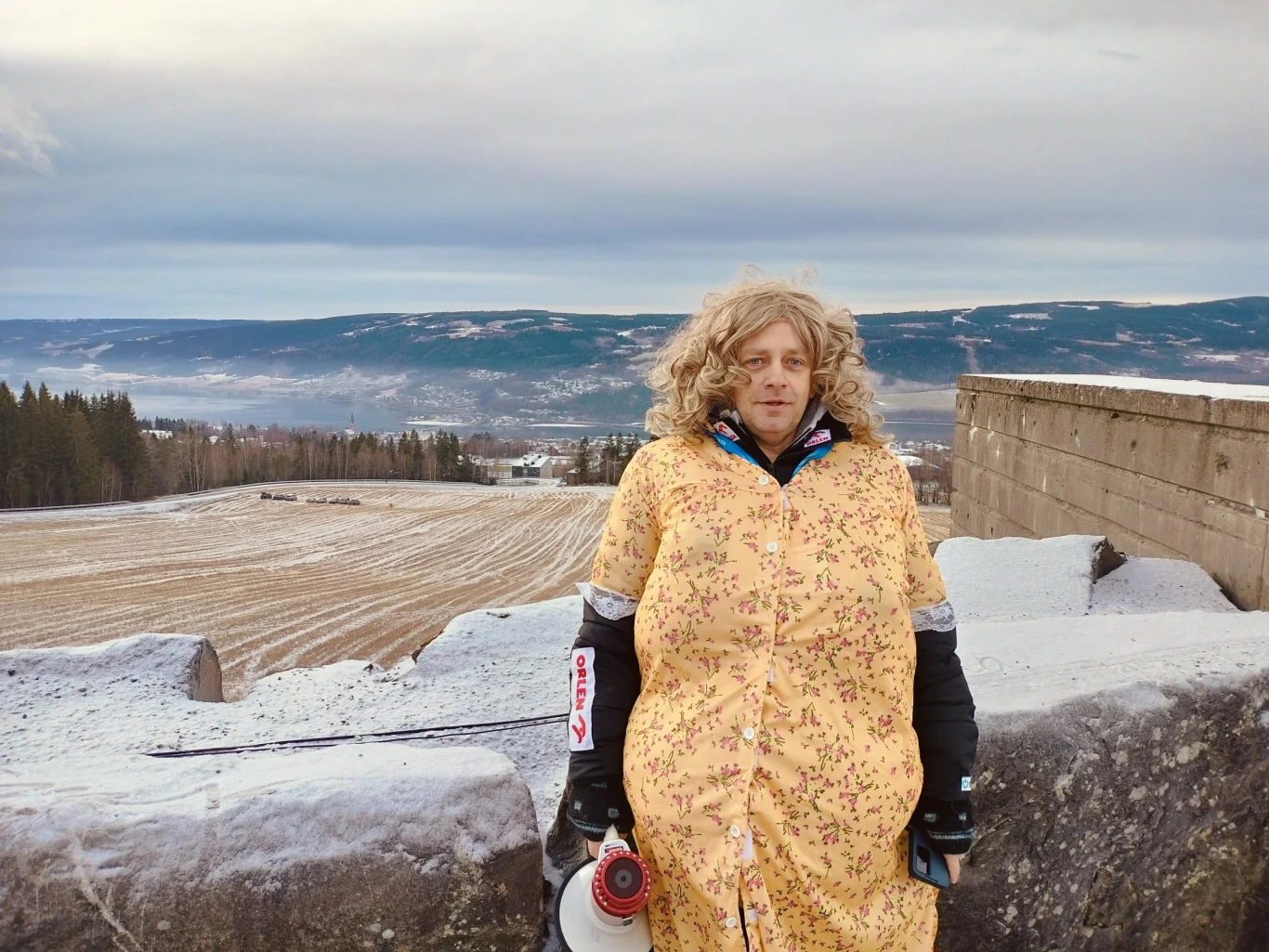 Mężczyzna w żółtej sukience z wzorem kwiatowym i peruce stoi na betonowej konstrukcji na tle zaśnieżonego pola oraz gór i lasu w oddali. Trzyma megafon i ma na sobie rękawice sportowe.
