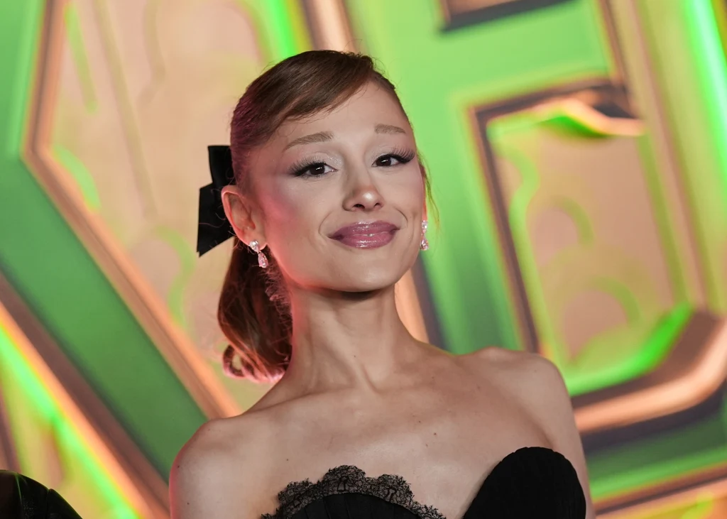 "Wicked: For Good": Ariana Grande w roli Glindy Kobieta z uśmiechem na twarzy ubrana w elegancką, czarną sukienkę, stojąca na tle wyrazistego zielono-żółtego wzoru, włosy spięte z dużą czarną kokardą, widoczne kolczyki.