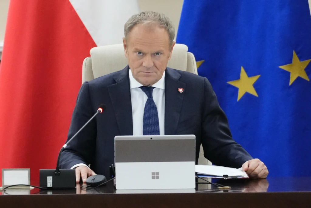 Premier Donald Tusk Mężczyzna w garniturze siedzi przy biurku z laptopem, obok mikrofon, za nim znajdują się flagi Polski i Unii Europejskiej.