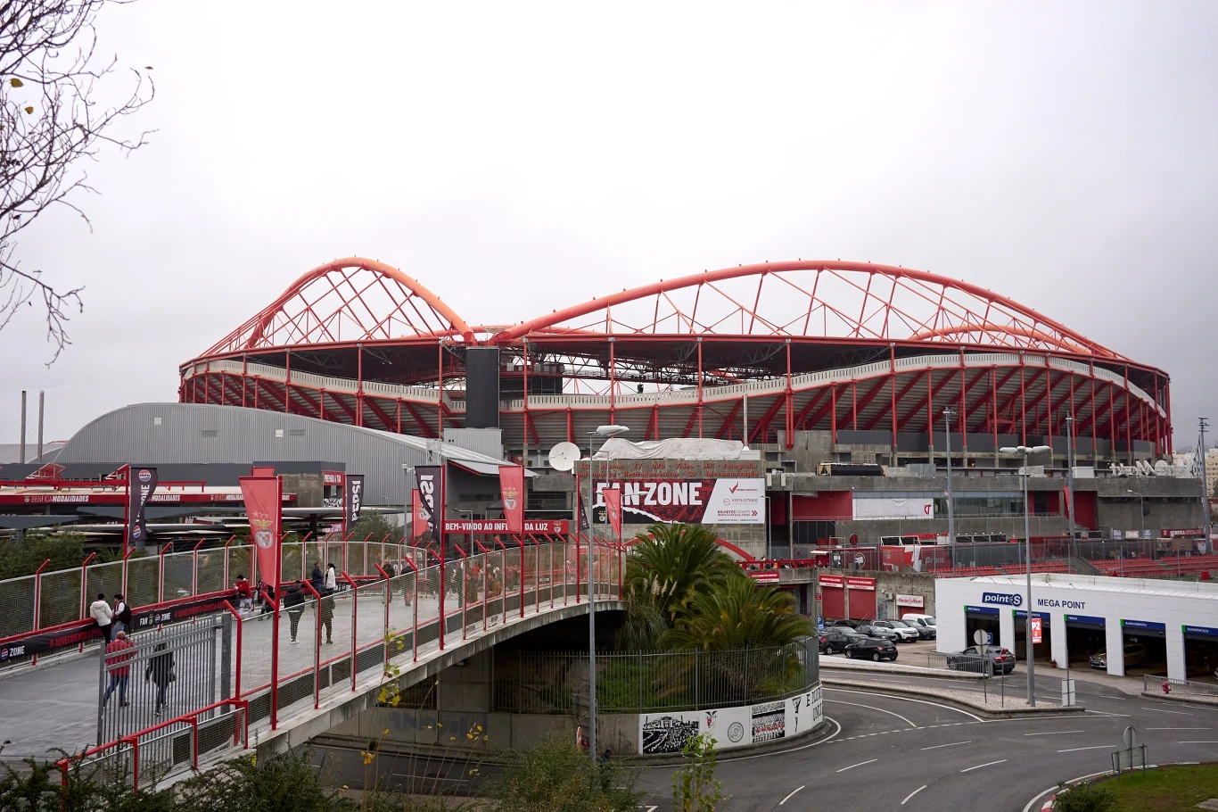 Nowoczesny stadion z charakterystyczną konstrukcją dachową w kolorze czerwonym, otoczony infrastrukturą miejską, drogami i przejściami dla pieszych. Przed stadionem widoczne elementy strefy kibica oraz samochody zaparkowane w pobliżu.