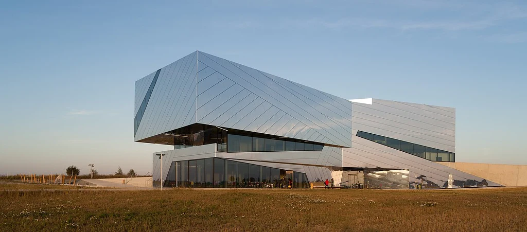 Muzeum w Schöningen, gdzie eksponuje się znaleziska sprzed 300 tys. lat Nowoczesny budynek o geometrycznych, ukośnych kształtach z dużymi przeszklonymi powierzchniami, wykonany z jasnych materiałów, otoczony rozległym trawnikiem, na tle jasnego nieba.