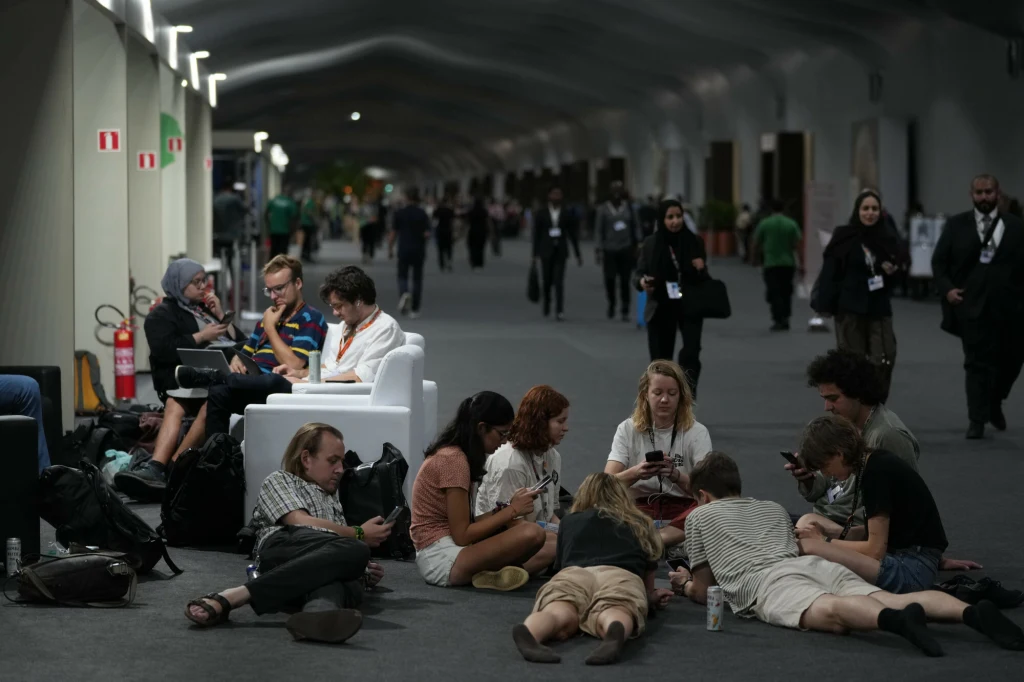 Ekologowie czekający na efekt obrad szczytu klimatycznego w Belem Grupa młodych ludzi siedzących na podłodze i na sofie w szerokim korytarzu, większość z nich zajęta korzystaniem ze smartfonów, w tle widoczne osoby spacerujące po hali.